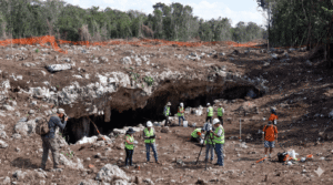 Group of construction workers and surveyors in safety vests near a rocky excavation with a large cave opening in the foreground.