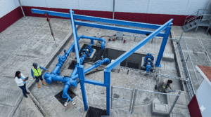 Aerial view of a blue piping system with pumps and valves in an industrial yard, workers in safety vests inspecting equipment.
